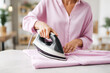 © Nadezda Ledyaeva - Woman in pink blouse using a steam iron on a light pink shirt, demonstrating the ironing process in a bright, modern home environment with soft natural light