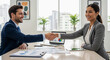 © dubogray1985 - Two business people shaking hands across a table in a modern office environment, with documents, charts, and a laptop visible.