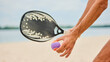 © Lustre Art Group  - Close up of male hand holding paddle racket and small ball on sandy beach. Concept of summer sport, lifestyle, equipment, outdoor leisure and playful emotional anticipation.