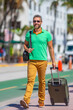 A tourist walks through the summer city streets. A man enjoys his summer vacation in a tropical summer city. Traveling with a suitcase. A full-length portrait of a traveler on an urban summer