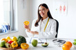 © Home-stock - Portrait of happy woman nutritionist holding orange fruit and smiling at camera, working at weight loss clinic, making meal plan for client