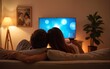 © Joseph - Back view of young couple enjoying themselves and watching tv on the sofa in the living room. Young man and woman relaxing and drinking red wine in the new flat. Man is hugging his girlfriend.