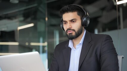 Thoughtful businessman in formal suit and headphones works on a laptop sitting in a business office. Serious coder developer or IT specialist is programming, busy with a project on computer. Close up
