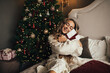 © polinaloves - Young beautiful woman with long curly hair sitting on a bed near Christmas tree, holding present.