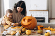 © sonyachny - Happy mother and cute baby son carving pumpkin on table with ghosts, spiders and bats decoration. Family cutting halloween pumpkin, making a jack o lantern. Halloween preparation