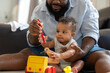 © Supachai - African American dad helps his baby play with colorful wooden toys while sitting together on the floor in home.