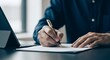 © ColorNest - Close up of a business professional in a suit writing with a pen on a document at a desk with a tablet