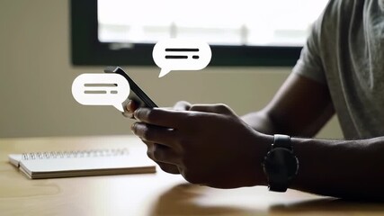Messaging: A Black man in his 30s replies to chat bubbles on his mobile phone indoors while seated at a wooden table, wearing a smart watch.