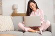 © Pixel-Shot - Young woman in striped pajamas working with laptop on sofa at home