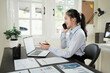 © Stella 16 - Asian businesswoman talking on cell phone while analyzing documents and working on laptop in office.
