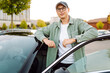© maxbelchenko - A young man wearing glasses and holding a phone stands next to a car in a parking lot. The man enjoys the sunny day with his phone next to a dark car. Conceptual image: transportation, leisure.