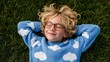 © iuricazac - A young child wearing glasses and a blue sweater, lying on grass with arms behind head, smiling contentedly.