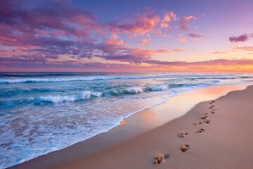 Wall Mural - Pink sunset sky over ocean waves lapping a sandy beach with footprints. Perfect for travel, vacation, nature, or inspirational backgrounds.