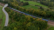 © VladyslavShcherbakov - Aerial view of passenger train moving through green countryside with winding road and fields, modern rail transport integrated into rural landscape and sustainable mobility concept.