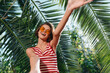 © SHOTPRIME STUDIO - A cheerful woman wearing a striped top and sunglasses takes a sunny selfie under palm fronds, smiling broadly. The vibrant outdoors evokes carefree summer vibes and a lively fashion moment.