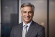 © The 2R Artificiality - Studio portrait of confident mature businessman wearing grey suit and tie, smiling with blurred office background and warm natural lighting.