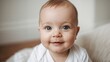 © The 2R Artificiality - adorable baby boy with bright blue eyes and a cheerful smile wearing a white onesie sitting on a soft textured blanket indoors with natural light