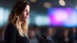 © Bussakon - A professional female speaker stands at a podium delivering a speech in a conference hall with a vibrant blurred screen in the background
