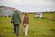 © Seventyfour - Senior Caucasian man and young adult Caucasian woman walking together on grassy field near small rural buildings, both seen from behind, spending time outdoors in countryside setting