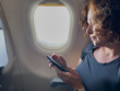 © Юлия Завалишина - Passenger woman is flying in plane. Girl using phone sitting by airplane window. Traveling female with smartphone inside plane enjoying flight. Woman using wifi connection on board. Traveling girl