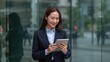 © The 2R Artificiality - Professional businesswoman in a suit using a digital tablet outdoors, smiling while standing near a glass building in a modern urban environment, showcasing corporate technology use.