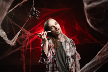 Creepy man with scary Halloween zombie makeup trapped spider webs listening voice message on smartphone. Dead guy in cob web on red bloody background.
