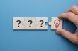 © MediaStockAI - A hand placing the final puzzle piece featuring a red lightbulb icon onto the puzzle, representing a solution or idea, adjacent to question marks on a light wood background, over a vibrant blue...