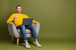 © deagreez - Young man in casual yellow shirt using laptop while sitting in chair with thumbs up on green background