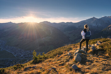 Wall Mural - Young woman with backpack standing on grassy mountain ridge with view of valley with high mountain peaks during golden hour sunset in autumn. Hiking in Italy, Livigno. Girl in Alps in fall. Trekking