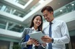 © miss irine - Asian business people in suits look at tablet. Woman holds blue folder, man wears tie. Colleagues smile, discuss project inside modern office atrium. Collaboration, teamwork, and success.