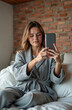 © Vadym - Young woman with rosacea takes selfie in bed. Female checks skin condition on phone screen. Woman wear bathrobe indoor, has facial redness and spots.