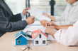 © doidam10 - House model on desk with blurred people discussing contract in background, concept of real estate investment, mortgage loan, home ownership, property business and financial agreement.