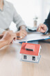 © doidam10 - House model on desk with blurred people discussing contract in background, concept of real estate investment, mortgage loan, home ownership, property business and financial agreement.