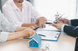 © doidam10 - House model on desk with blurred people discussing contract in background, concept of real estate investment, mortgage loan, home ownership, property business and financial agreement.