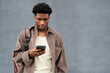 © Mediaphotos - Young Black man standing outdoors using smartphone, carrying shoulder bag, looking down at device with focused expression against plain gray background