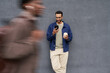 © Mediaphotos - Young Black man standing against gray wall holding coffee cup and smartphone, looking at screen and smiling, blurred figure of another young Black man walking past in foreground
