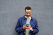 © Mediaphotos - Young Black man standing against plain gray wall using smartphone, wearing glasses and casual clothing, looking down at device with focused expression, holding phone with both hands
