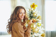 © Alliance - A woman with long, wavy brown hair and a warm smile holds a golden Christmas ornament in front of a decorated Christmas tree. Soft lighting illuminates the festive holiday atmosphere.