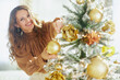 © Alliance - A close-up shows a smiling woman with wavy brown hair decorating a frosted Christmas tree with golden ornaments. Soft, natural light creates a warm and festive domestic scene.