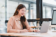 © kenchiro168 - Young woman smiling at laptop in modern coworking space, productive remote worker enjoying creative focus and bright natural light