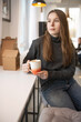 © shymar27 - Young woman with long brown hair, dressed in a cozy gray sweater, is sitting comfortably on a sofa in a modern cafe, showcasing a relaxed and inviting atmosphere
