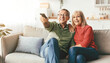 © Prostock-studio - Happy Senior Spouses Watching TV Pointing Television Remote Control Sitting And Hugging On Sofa At Home. Mature Couple Enjoying Favorite Movie Together On Weekend. Retired Family Leisure