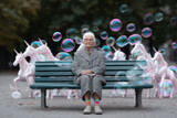 Elderly woman on a park bench, surrounded by bubbles and ethereal unicorn figures. A visual metaphor for dreams, memory, and imagination in aging. Conceptual  whimsical.