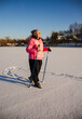 © StockMediaSeller - Elderly woman walking with trekking poles on a snowy field during sunny winter day. She wears a pink jacket and smiles, enjoying outdoor exercise and fresh air in the cold season.