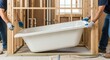 © MT - Two male construction workers carefully installing a new white bathtub into a framed bathroom during a home renovation project, with exposed plumbing visible.