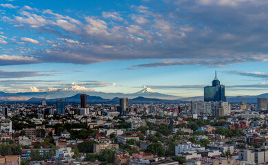 Wall Mural - A city skyline with a cloudy sky in the background. The city is full of tall buildings and trees. Panoramic view of Mexico City, snow-covered volcanoes Popocatepetl and Iztaccihuatl