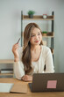 © WMSTUDIO - Thoughtful professional woman in light sweater sitting at desk with laptop, holding pen, contemplating ideas in modern workspace with greenery and books