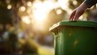 © rehan - Someone's hand gently touches the lid of a green garbage bin, bathed in warm sunlight from a bright, blurred background, evoking a sense of everyday life and outdoor activities.