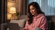 © Ashura - A young woman in a pink hoodie smiles while working on her laptop in a cozy, dimly lit living room.