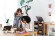 © Studio Marmellata - Two young women collaborate on a laptop at a wooden desk, surrounded by plants and office supplies, in a bright, modern workspace.